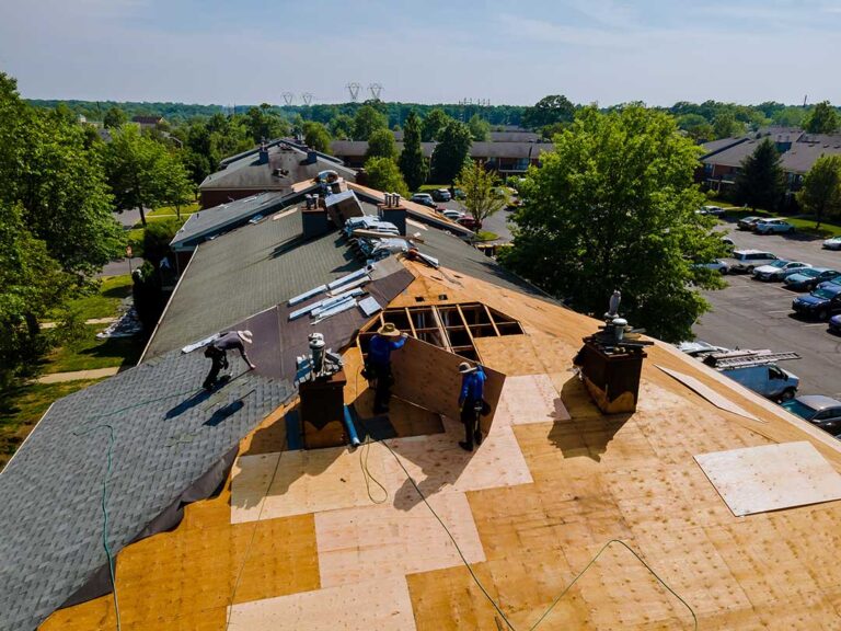 A person wearing safety gear uses a long-handled roof rake to clear leaves from a house roof.
